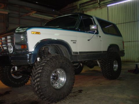 1986 Ford Bronco - Three-quarter front view of a lifted white and teal two-tone Ford Bronco with removable hardtop, parked inside a metal shop building. It si…