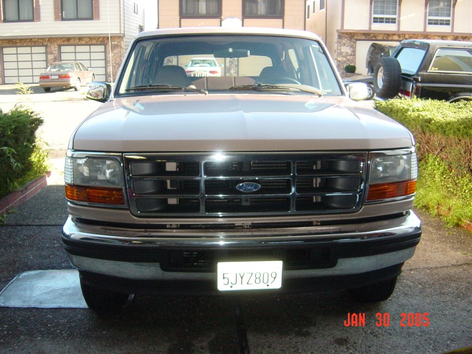 Front view of a tan Ford Bronco with a chrome grille. Another Bronco is parked to the right