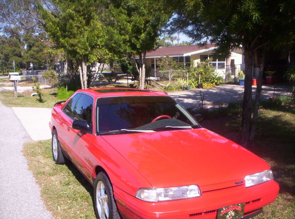Red Mazda coupe with sunroof parked on grass beside driveway, front 3/4 view