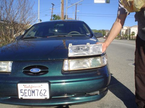 1993 Ford Taurus - A person holds a replacement headlight assembly next to the driver's side headlight of a dark green 1992 Ford Taurus.