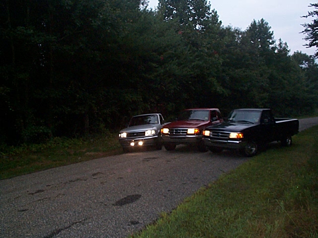 Three pickups parked on a road at dusk with headlights on, silver, red, and black.