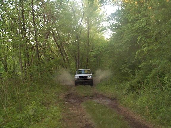 Silver pickup splashing through mud on a wooded dirt trail, front view