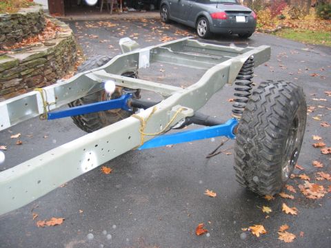 1978 Ford Bronco - A bare, freshly painted light-green vehicle frame sits on a wet paved driveway, fitted with a solid axle, blue-painted radius arms, coil sp…