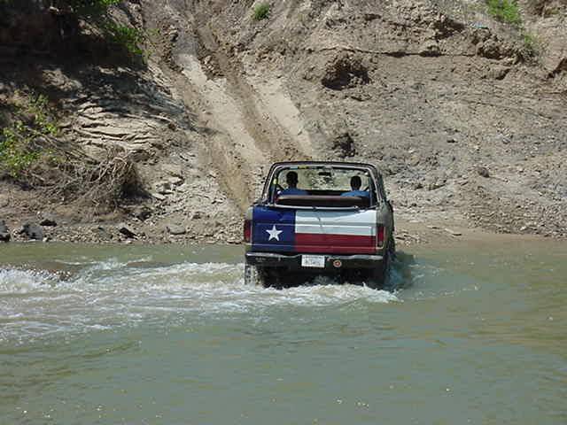 Topless Ford Bronco with Texas flag tailgate fording a muddy river