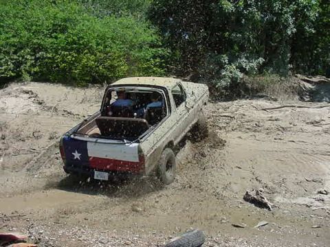1978 Ford Bronco - Tan topless lifted Ford 4x4 with a Texas flag tailgate blasting through a shallow mud pit, spraying muck off the rear tire. Shot from behin…