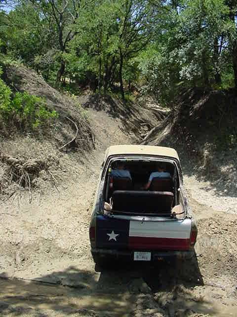 1978 Ford Bronco - Rear view of a topless full-size Ford Bronco with a tan roll bar hoop and a Texas-flag-painted tailgate, working up a sandy washed-out trai…
