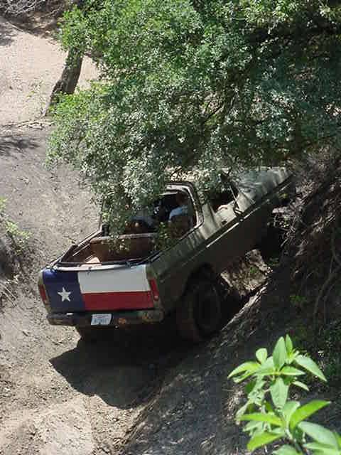 1978 Ford Bronco - Overhead rear view of an olive-drab open-top 4x4 with the roof removed, working down a steep rutted dirt trail under overhanging oak branch…