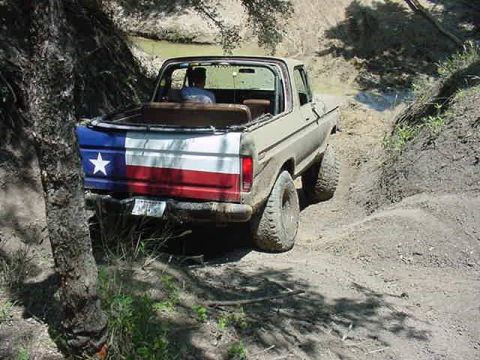 1978 Ford Bronco - Rear three-quarter view of an olive-green topless full-size Ford Bronco with a Texas-flag-painted tailgate easing down a rutted dirt trail…