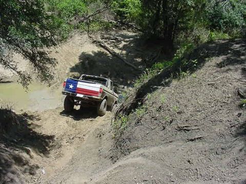 1978 Ford Bronco - Rear view from above of a tan topless Ford Bronco with a Texas flag painted across its tailgate, climbing out of a muddy creek crossing ont…