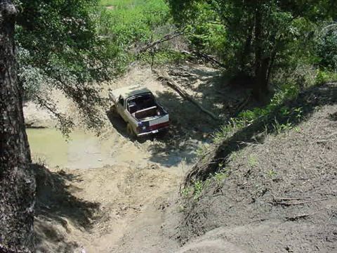 1978 Ford Bronco - Elevated view down into a muddy water hole where a light-colored open-top full-size Bronco with a white roll bar works through churned brow…