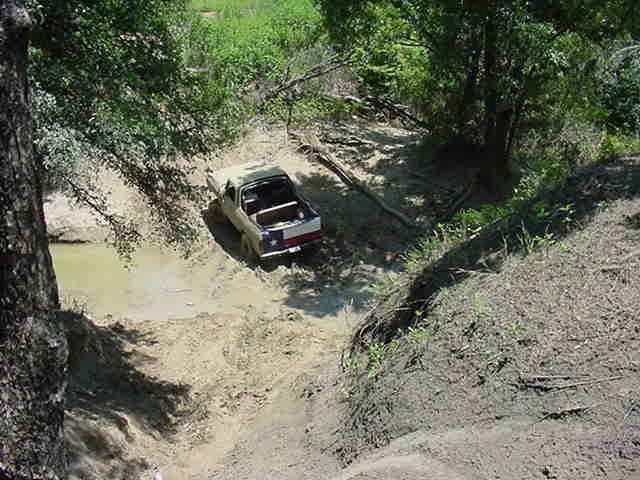 Open-top Bronco with roll bar crossing a muddy water hole on a wooded trail, viewed from above