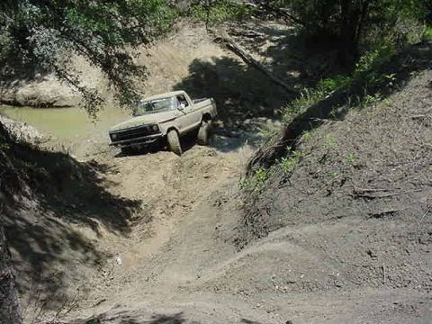 1978 Ford Bronco - Shot from above a dirt bank, a tan topless Ford Bronco with the roof removed noses out of a muddy water hole at the bottom of a steep sand…