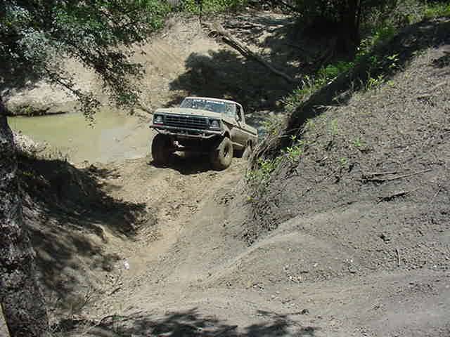 Muddy late-1970s Ford Bronco climbing out of a rutted gully next to a water hole