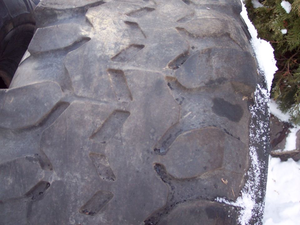 Close-up of a large, aggressive tire tread pattern with snow on the sidewall and a bush in the background. Supermotors.net logo visible