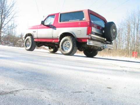 1995 Ford Bronco - This two-tone red and silver SUV, likely a 1995 Ford Bronco, is seen from the rear on a snowy road. A spare tire is mounted on the tailgate.