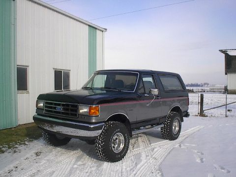 1989 Ford Bronco - A two-tone Ford Bronco with off-road tires sits in a snowy landscape next to a building.