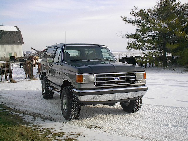 Dark gray Ford Bronco with chrome grille and bumper, lifted on off-road tires, parked on a snowy dirt road