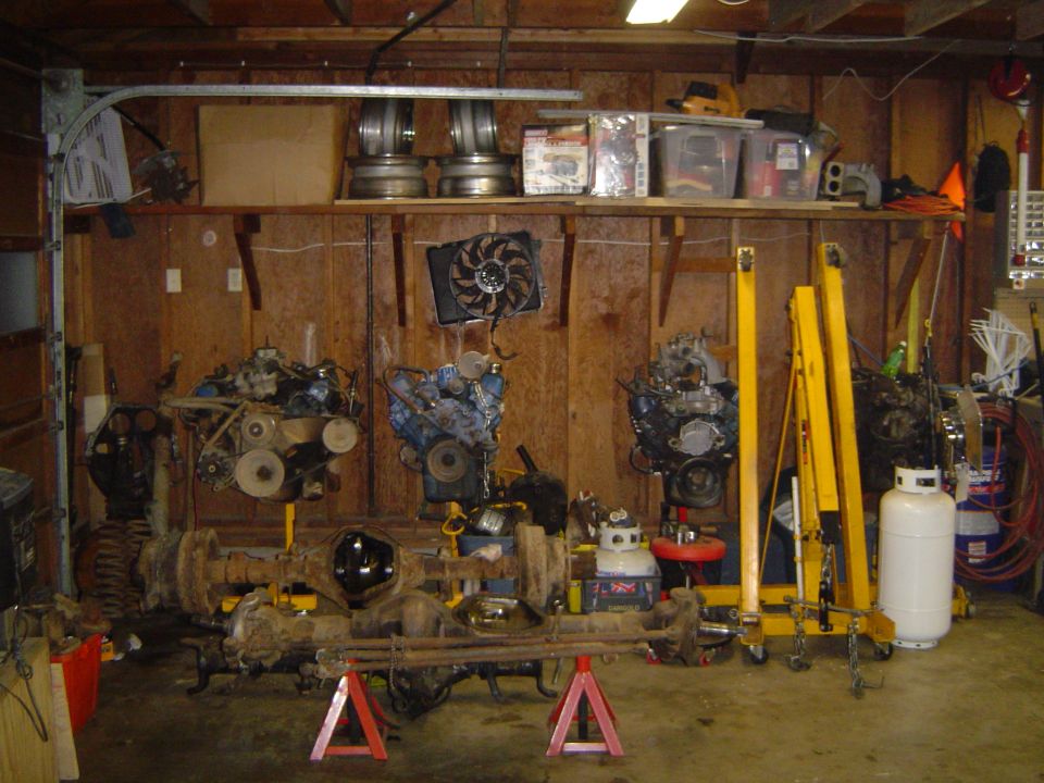 Garage with three V8 engines on stands and a rusty solid axle on jack stands