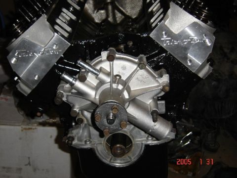 1990 Ford Bronco - Front view of a black V8 short block on a stand with a cast aluminum timing cover and crank hub installed, flanked by two bare aluminum cyl…