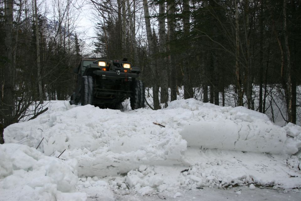 Olive green all-terrain vehicle with tracks, driving through deep snow in a forest
