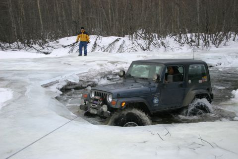 2003 Jeep TJ - A Jeep Wrangler navigates through a partially frozen body of water with a person observing from the snowy bank.