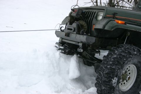 2003 Jeep TJ - A green 2003 Jeep TJ's front bumper and large tire are partially buried in snow, with a winch line extending from the vehicle.