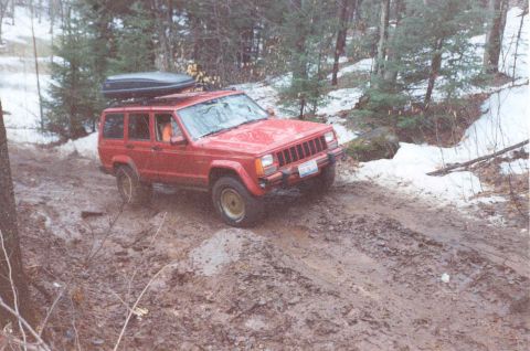 1995 Ford Bronco - A red Jeep Cherokee XJ navigates a challenging muddy path through a snowy forest.