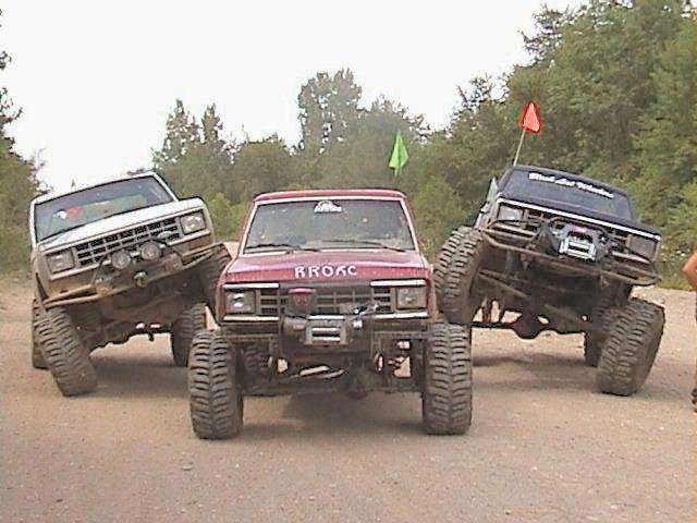 Three lifted 4x4 trucks posed front-on, outer ones tilted onto center red winch-equipped SUV on gravel.