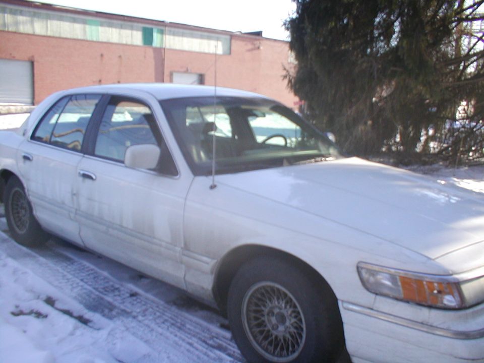 White sedan parked in snow, front quarter view. Building and trees in background