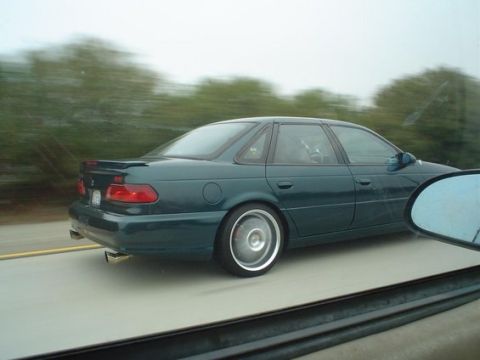 2002 Ford Taurus - This dark green sedan, likely a 1998 model, is captured in motion on a highway, its side profile and custom wheels.