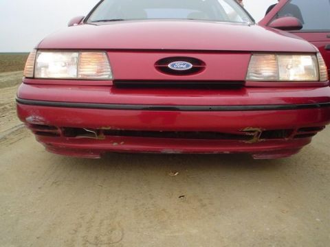 2002 Ford Taurus - The front of a red Ford sedan shows damage on the bumper and grille area. Another vehicle is partially visible.