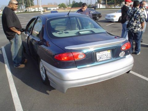 2002 Ford Taurus - A group of people stand around a dark blue sedan with a silver lower body kit and a spoiler in a parking lot.