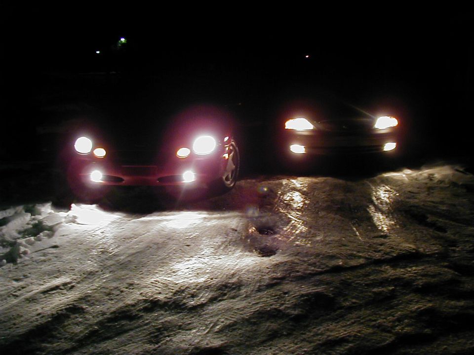 Two sedans at night with headlights on, parked on snow-covered ground