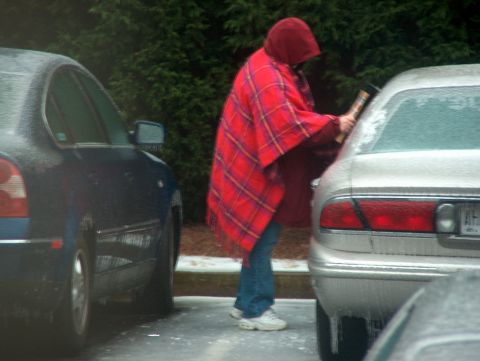 1998 Ford Taurus - A person in a red plaid poncho scrapes ice from the rear window of a silver car parked next to a blue car.