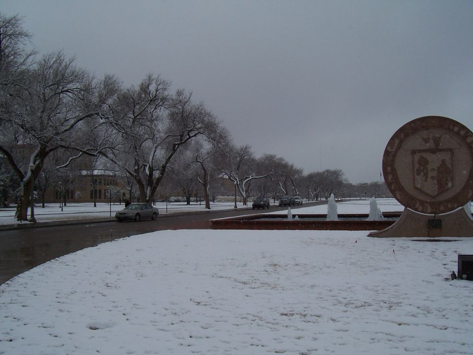 Silver sedan parked on a snow-covered campus mall beside a round stone seal monument
