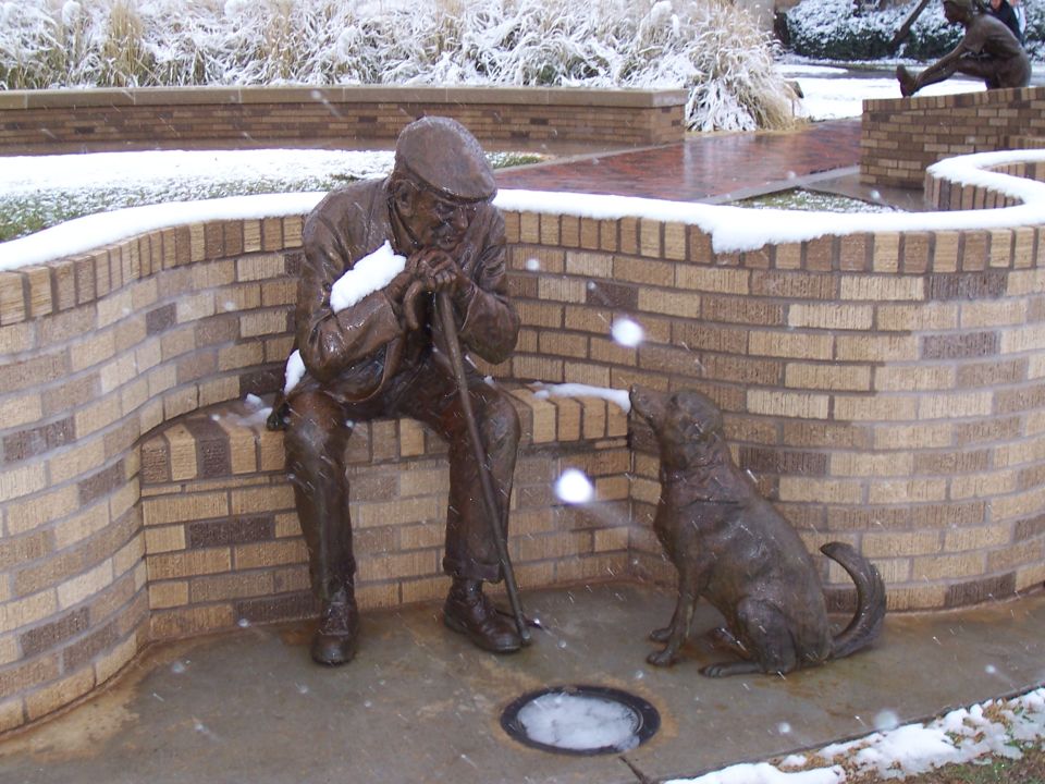 Snow falling on a bronze statue of a seated old man with a cane and a bronze dog