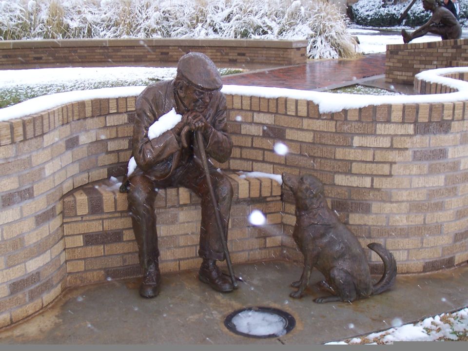 Bronze statue of an old man on a brick bench facing a bronze dog during snowfall