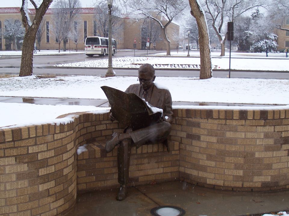 Bronze statue of a man reading a newspaper with a white transit bus passing behind in the snow