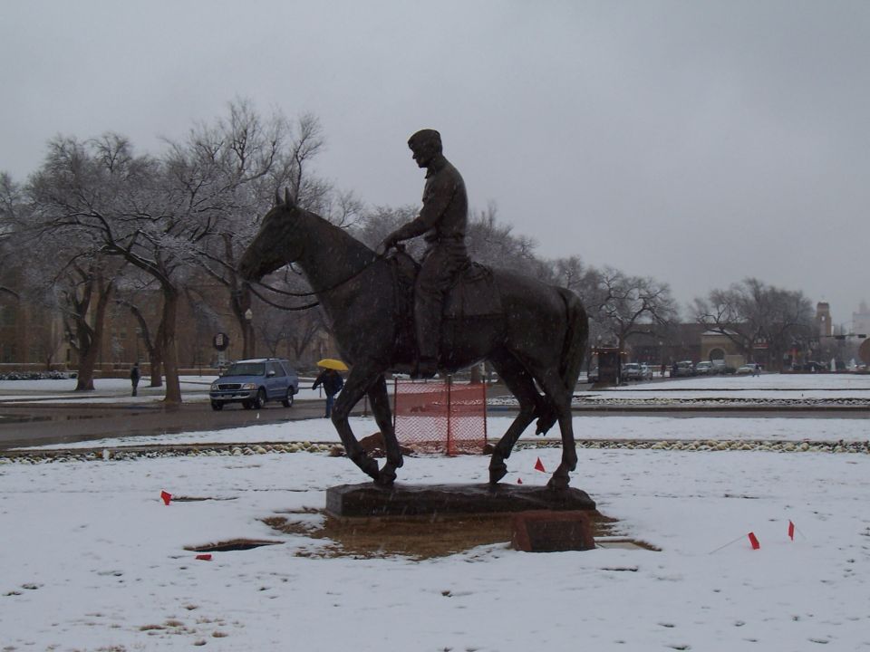 Bronze horse-and-rider statue in a snowy campus plaza with orange fencing and a parked SUV