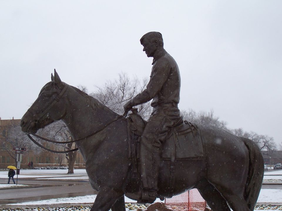 Bronze statue of a man on horseback during a snowfall on a campus lawn