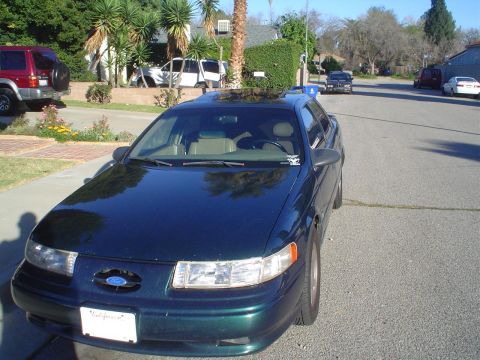 1993 Ford Taurus - A dark green sedan is parked on the side of a street in a suburban neighborhood.