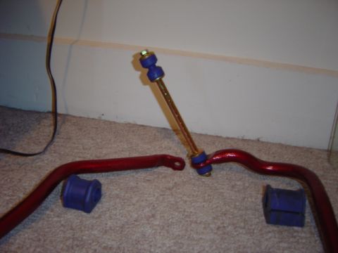 1998 Ford Taurus - Two red powder-coated sway bars with blue polyurethane bushings and a matching end link, laid out on tan carpet against a wall.