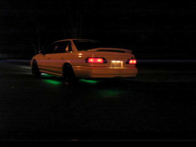 Yellow 1995 Ford Taurus sedan with green underglow and a spoiler, seen from the rear at night