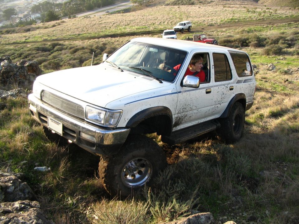 Muddy white lifted Toyota 4Runner front quarter with driver in red on grassy slope