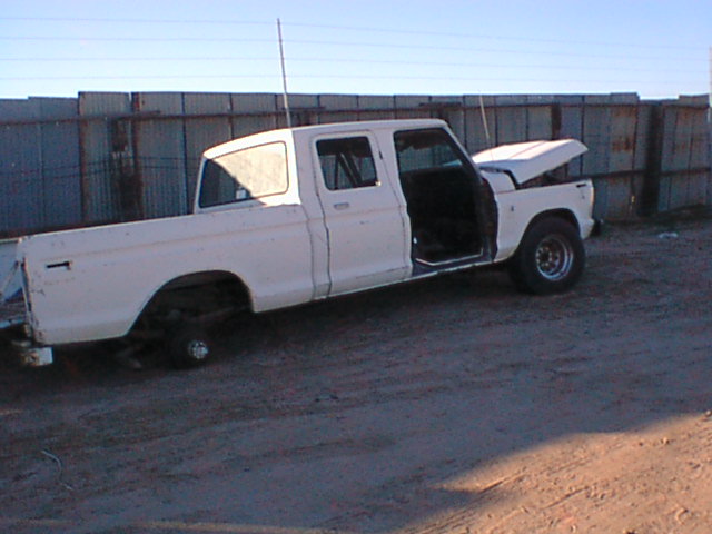 White Ford crew cab pickup on dirt lot with hood up, rear door open, and front wheel missing