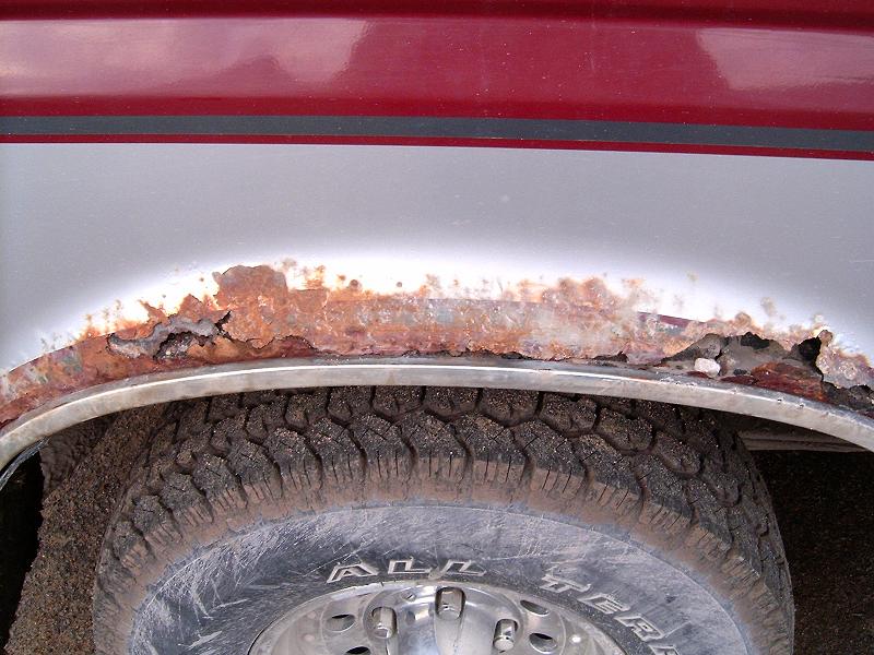 Heavy rust perforation along a red and white truck's rear wheel arch above all-terrain tire