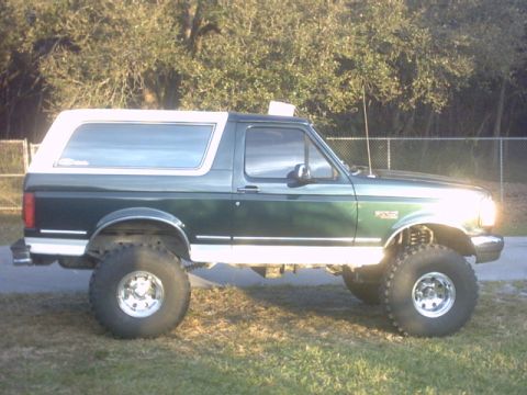 1992 Ford Bronco - This lifted 1992 Ford Bronco is shown from the side, parked on grass with a fence and trees in the background.