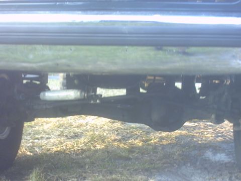 1992 Ford Bronco - Low ground-level view under the side of a truck parked on dry grass, showing the boxed frame rail, a muffler or resonator canister, exhaust…