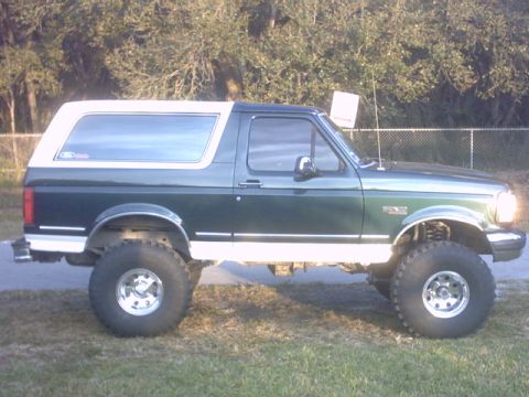 1992 Ford Bronco - Passenger-side profile of a lifted full-size Ford Bronco in dark green with a white removable hardtop and white lower rocker trim, on polis…