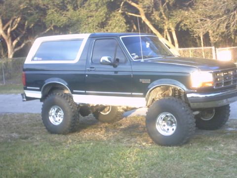 1992 Ford Bronco - Lifted dark green full-size Ford Bronco with a white removable hardtop, photographed from the passenger side on grass beside a chain-link f…